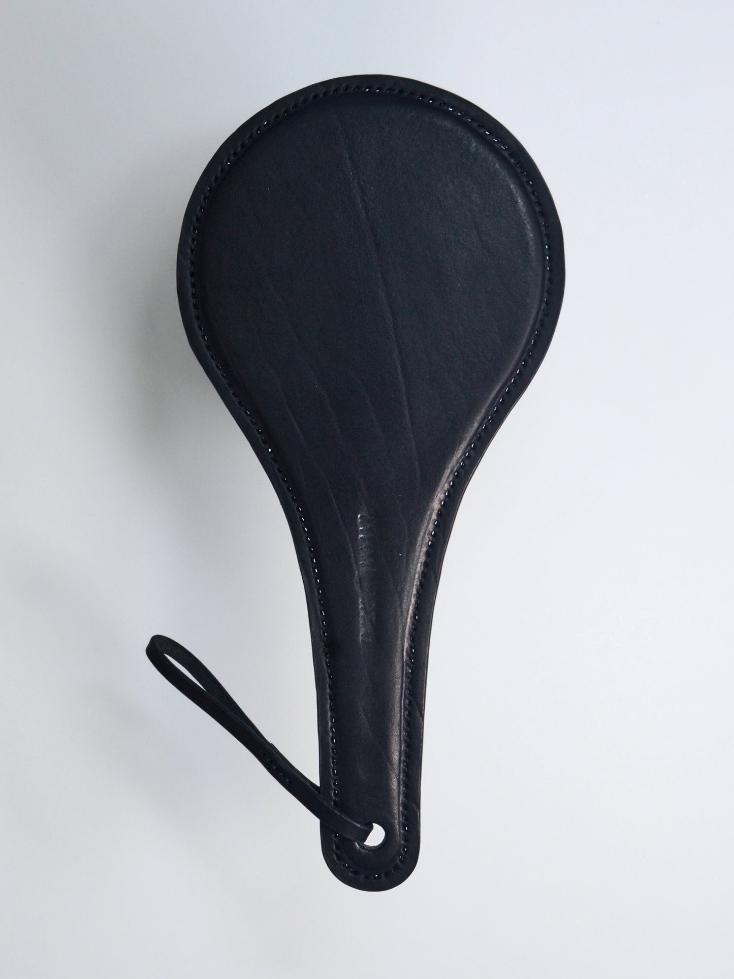 A black leather paddle with a thin strap with the words IM.MORTAL embossed in the middle of the handle, showcased against a white background.