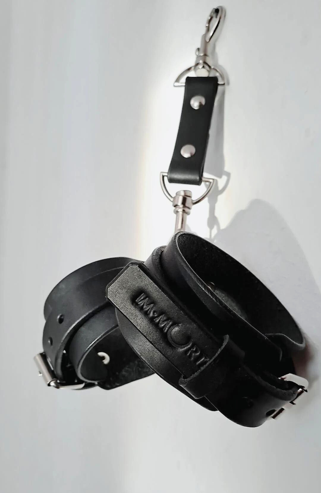 A pair of black leather cuffs, one designed for the wrists with metal rivets and a small chain attached for a hog-tie setup.