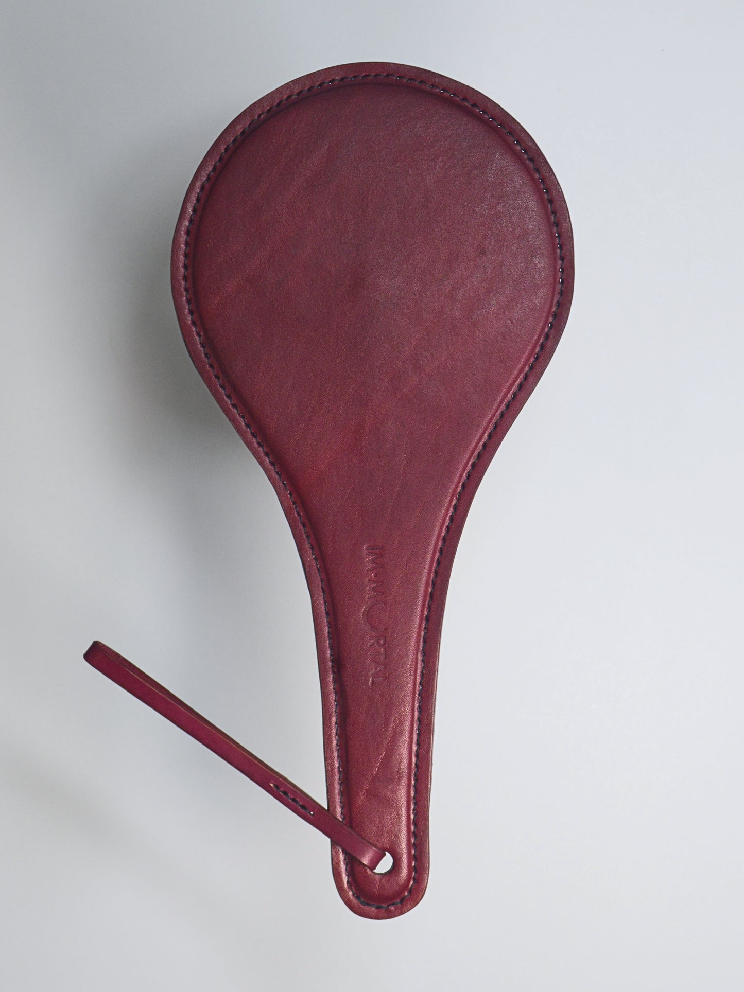 A burgundy leather paddle with a thin strap  with the words IM.MORTAL embossed in the middle of the handle, showcased against a white background.