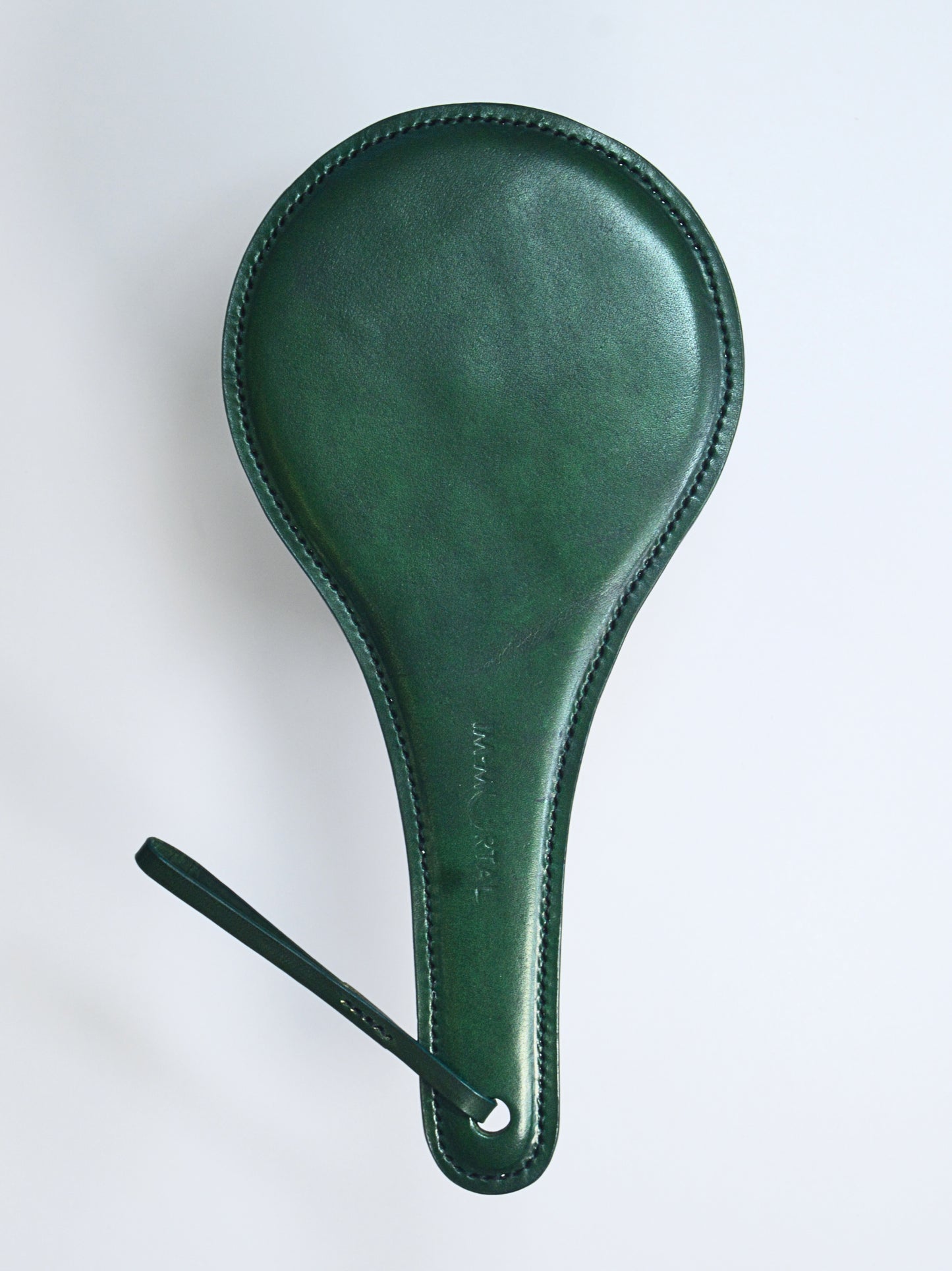 A green leather paddle with a thin strap  with the words IM.MORTAL embossed in the middle of the handle, showcased against a white background.