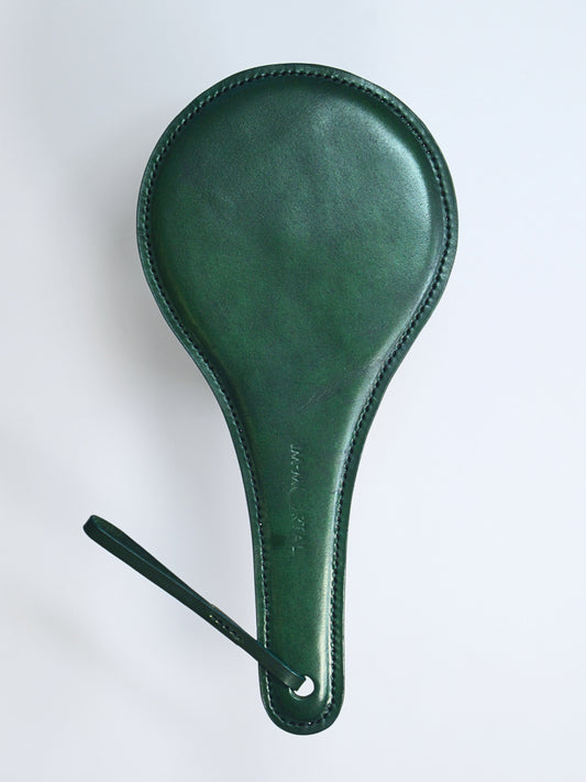 A green leather paddle with a thin strap  with the words IM.MORTAL embossed in the middle of the handle, showcased against a white background.