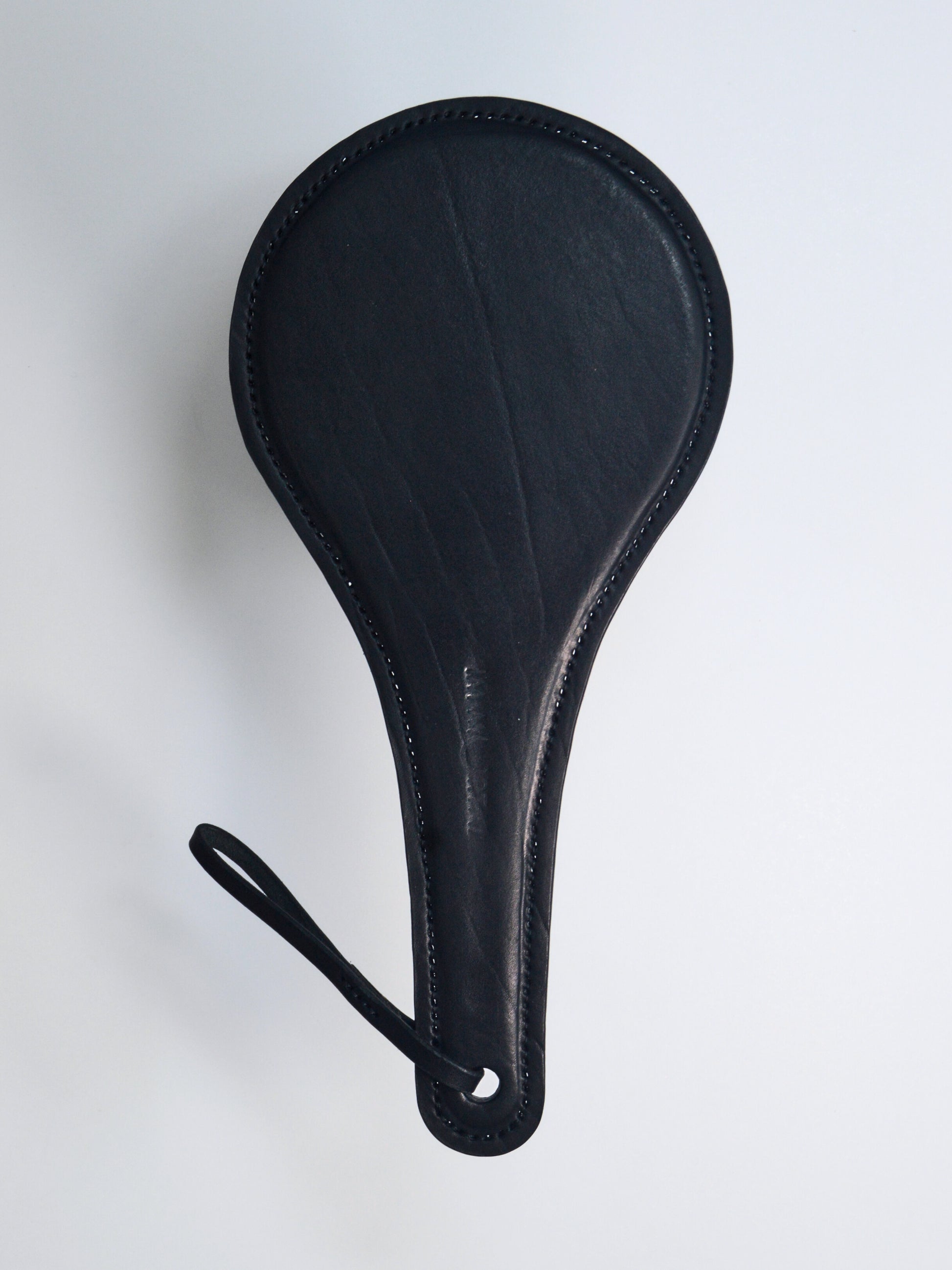 A black leather paddle with a thin strap  with the words IM.MORTAL embossed in the middle of the handle, showcased against a white background.