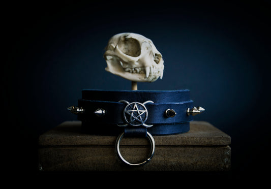 A dark-colored leather collar with a metal buckle and a silver-colored pendant featuring a pentagram design, displayed on a surface with a skull in the background.