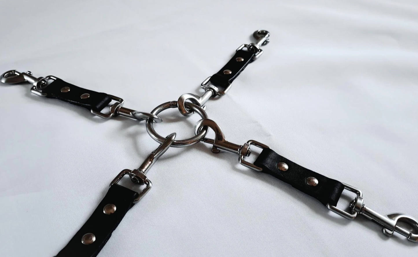 Four black leather straps with metal clasps are connected by a central metal ring (multi-functional leather hog-tie), arranged on a white surface.