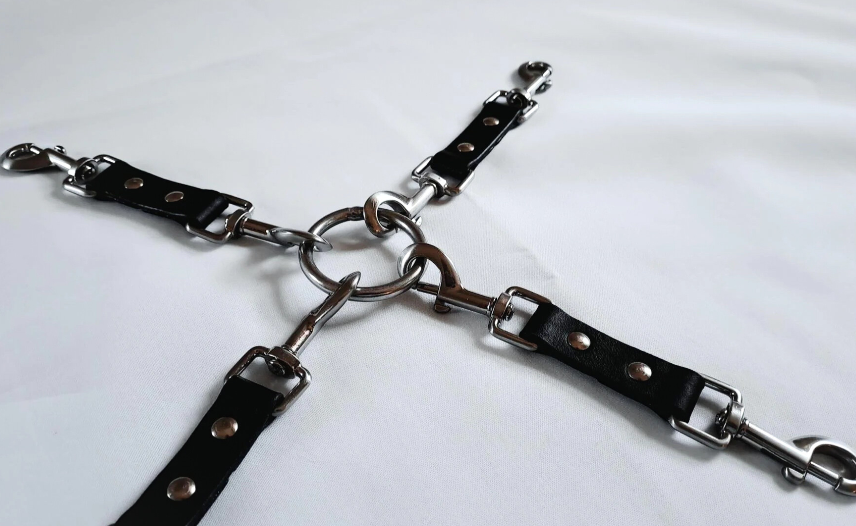 Four black leather straps with metal clasps are connected by a central metal ring (multi-functional leather hog-tie), arranged on a white surface.